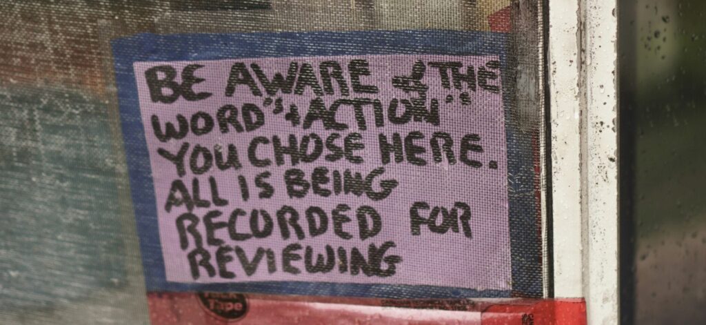 Photograph of a sign posted on a screen door, saying "Be Aware of the Word's + Action's you chose here. All is being recorded for reviewing"