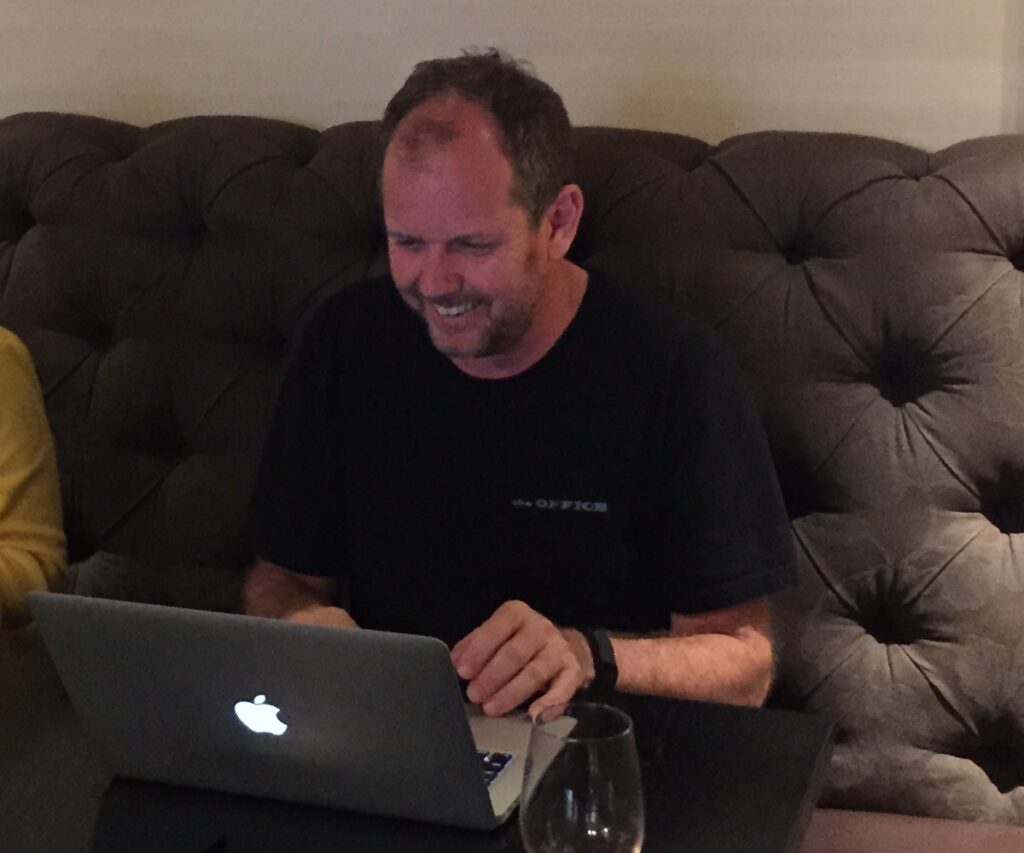 Photograph of Andrew Nash, sitting at a table working on his laptop, his characteristic smile on his face.